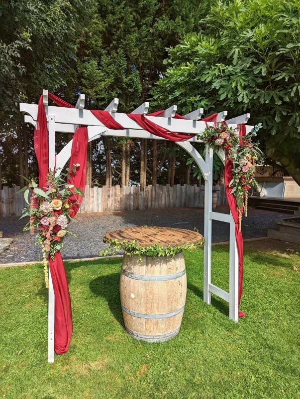 Arche de cérémonie de mariage champêtre ornée de draperies rouges et de compositions florales de roses, dahlias et eucalyptus à Génissac.