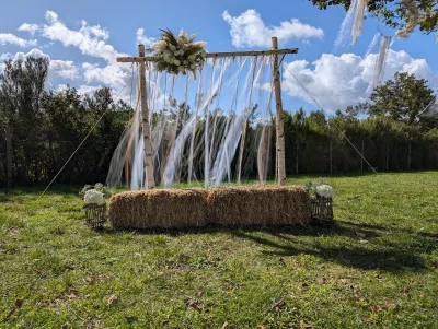 Arche de cérémonie bohème en bois de bouleau, ornée de pampas, roses blanches, feuillage et rubans flottants, avec des ballots de foin rustiques.