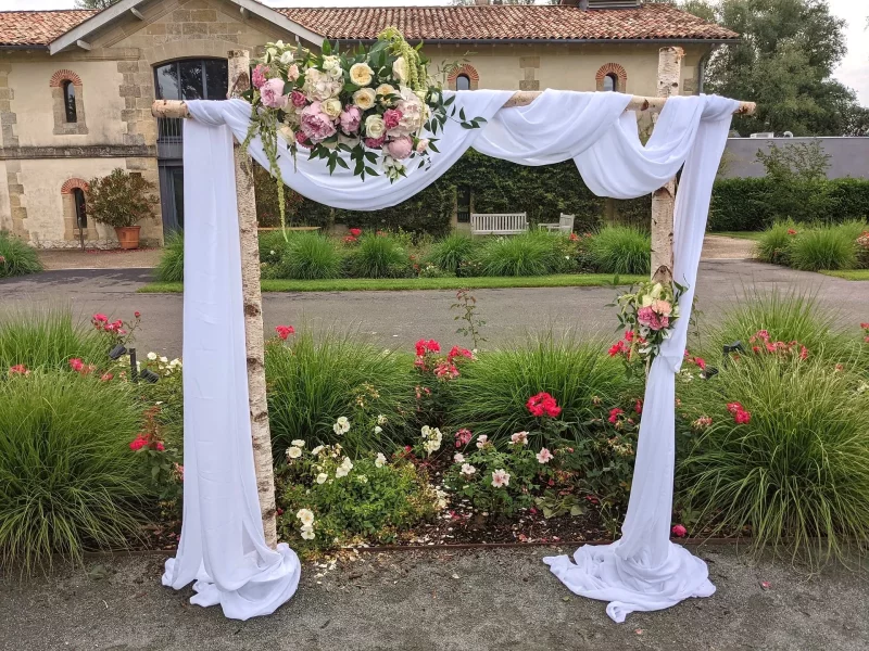 Arche de mariage classique en bouleau et tissu blanc, décorée de compositions florales roses et blanches pour la cérémonie.