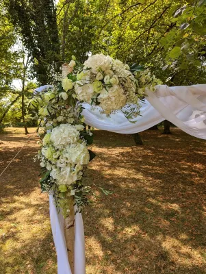 Arche florale décorée d'hortensias blancs, de renoncules crème et de feuillage vert varié, drapée de tissu, cérémonie extérieure.