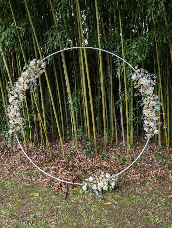 Arche de mariage circulaire moderne décorée de roses poudrées, eustoma et feuillage argenté devant une bambouseraie.