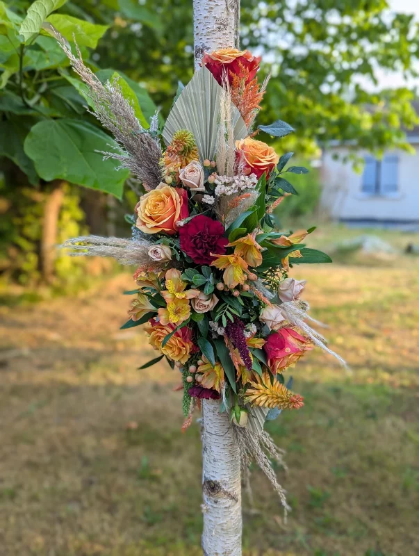Arche florale de mariage bohème avec roses orange, bordeaux, pampa et palmier séché sur tronc d'arbre.