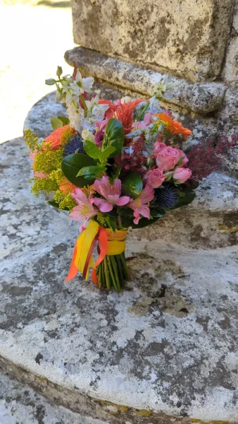 Bouquet de mariée champêtre vibrant pour un mariage en été, composé de roses corail, roses et jaunes, delphinium bleu et feuillage verdoyant.