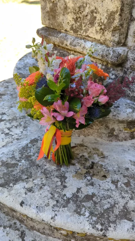 Bouquet de mariée champêtre vibrant pour un mariage en été, composé de roses corail, roses et jaunes, delphinium bleu et feuillage verdoyant.