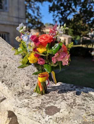 Bouquet de mariée champêtre vibrant pour un mariage en été, composé de roses corail, roses et jaunes, delphinium bleu et feuillage verdoyant.