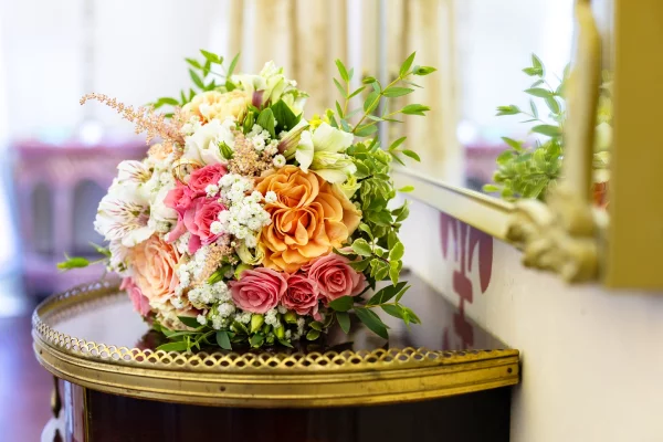 A garden-style bridal bouquet featuring peach-orange roses, coral roses, gypsophila, and verdant green foliage.