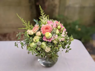 Bouquet de table champêtre et bohème avec gerbera rose, roses crème et feuillage varié dans un vase en verre.