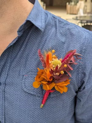 Sophisticated rustic buttonhole with dried orange, red, and yellow flowers, burgundy foliage on blue shirt.