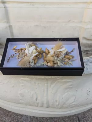 Boutonnière de mariage bohème avec des fleurs séchées couleur crème et des épis de pampas dans un coffret noir.
