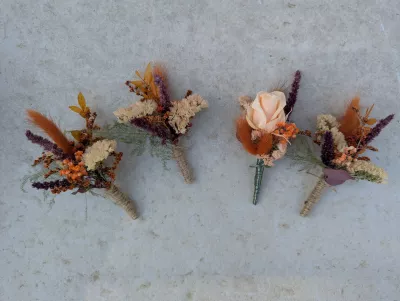 Four country-style buttonholes with dried flowers in orange, terracotta, burgundy, and a cream rose.