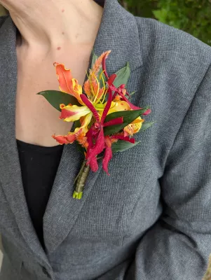 Boutonnière exotique vibrante avec gloriosa, petites orchidées vanda rouges et feuillage pour un mariage.