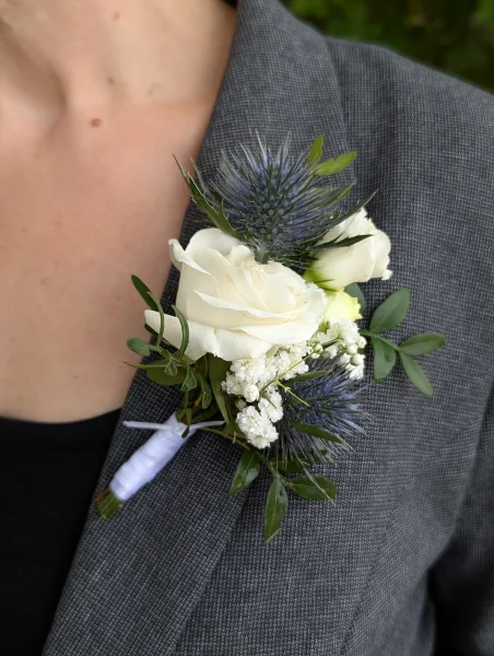 Boutonnière élégante pour le marié, composée d'une rose blanche, de chardons bleus et de gypsophile, pour un mariage champêtre.