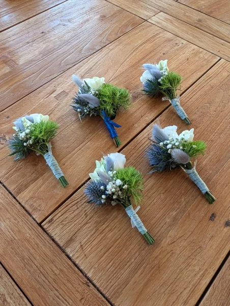 Boutonnières de mariage champêtres avec petites roses blanches, chardons bleus et feuillage pour les témoins.
