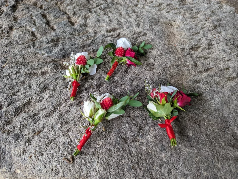 Boutonnière de marié champêtre avec roses blanches, fleur rouge pompon et feuillage d'eucalyptus, pour un mariage estival.