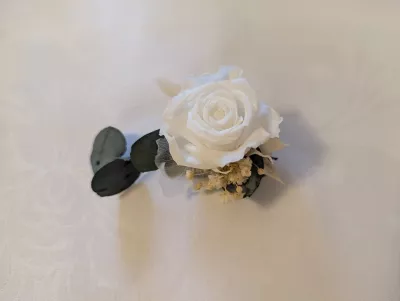 Boutonnière de mariage délicate avec une rose crème préservée, accompagnée de feuilles d'eucalyptus et de petites fleurs séchées.
