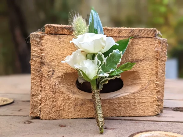 Boutonnière de mariage champêtre avec roses blanches, feuillage panaché et graminées, élégamment ficelée, sobre.