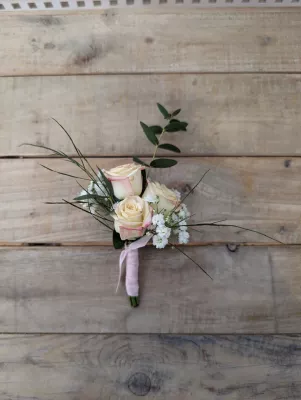 Boutonnière élégante style champêtre avec roses crème, gypsophile et feuillage vert, nouée d'un ruban rose pâle.