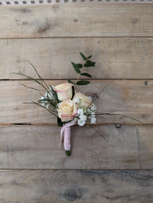 An elegant country-style boutonnière with cream roses, gypsophila and greenery, tied with a pale pink ribbon.