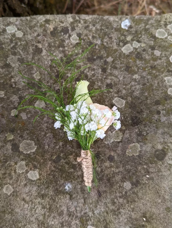 Boutonnière champêtre avec bouton de rose crème, gypsophile et feuillage fin, attachée avec de la ficelle de jute.