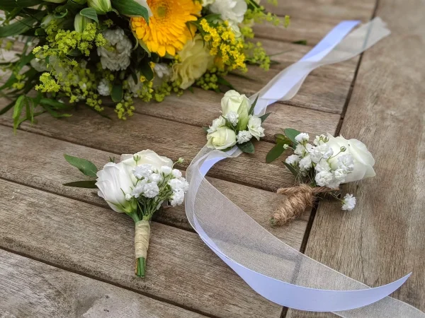 Boutonnières et bracelet floral de mariage classiques, confectionnés avec de délicates roses blanches et du gypsophile.