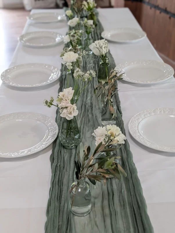 Centre de table champêtre pour mariage, avec des œillets blancs, roses pâles, branches d'olivier dans de petits vases en verre, ambiance naturelle.