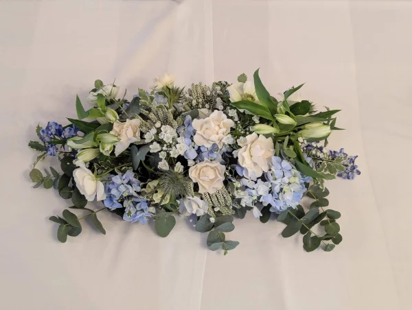 Composition florale de centre de table allongée classique avec roses, hortensias bleus, lys et eucalyptus pour mariage.