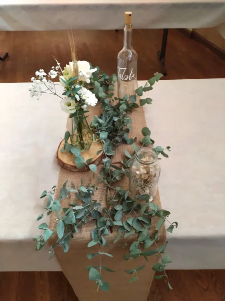 Centre de table mariage champêtre avec chemin de table en jute, eucalyptus, rose blanche, gypsophile et blé, style naturel.