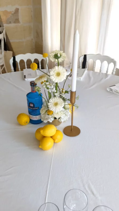Décoration de centre de table de mariage avec gerberas blancs, craspedias jaunes, et une bouteille bleue style limoncello