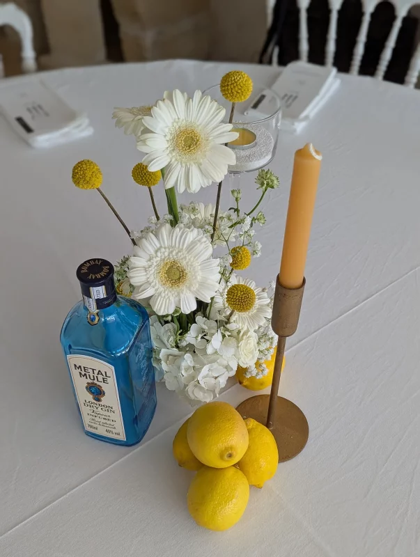 Décoration de centre de table de mariage avec gerberas blancs, craspedias jaunes, et une bouteille bleue style limoncello