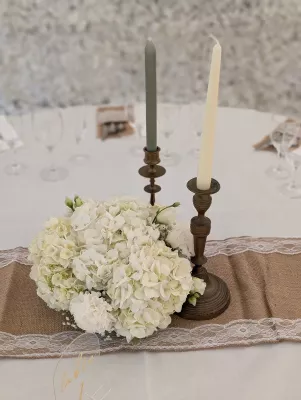Centre de table champêtre et bohème avec des hortensias et œillets blancs, gypsophile, sur un chemin de table en jute et dentelle, agrémenté de chandeliers vintage.
