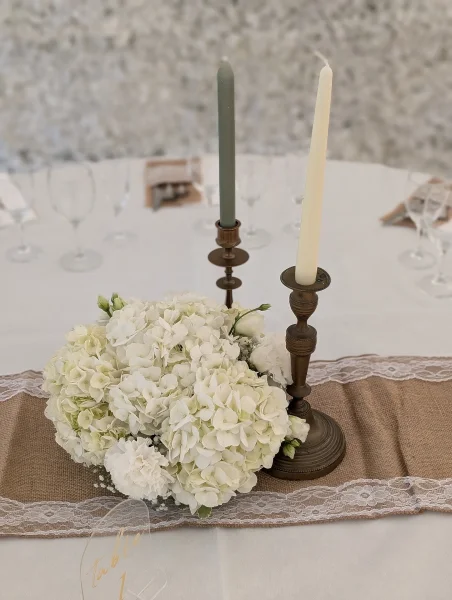Centre de table champêtre et bohème avec des hortensias et œillets blancs, gypsophile, sur un chemin de table en jute et dentelle, agrémenté de chandeliers vintage.