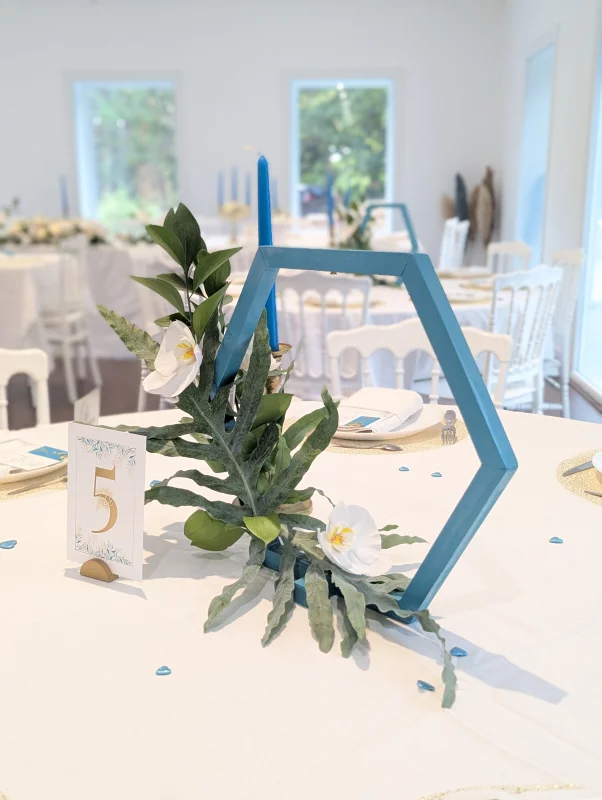 Sophisticated country wedding table centrepiece with white orchids, foliage, and blue hexagonal structure.