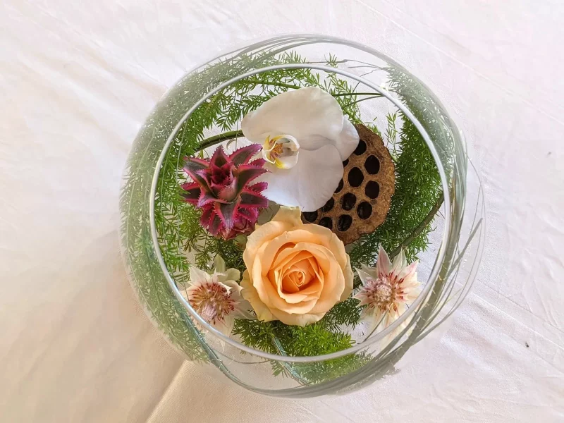 Vue plongeante d'un centre de table exotique avec orchidée blanche, rose pêche, guzmania et lotus séché