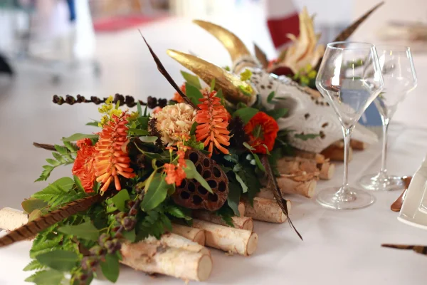 Long centre de table bohème avec renoncules, œillets, feuillage et cornes dorées sur bûchettes de bois.