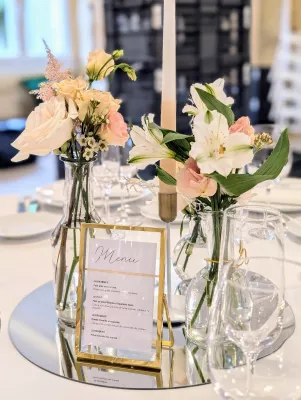 Centre de table de mariage classique avec roses, alstroemères, lisianthus dans des vases, bougie et menu sur miroir.