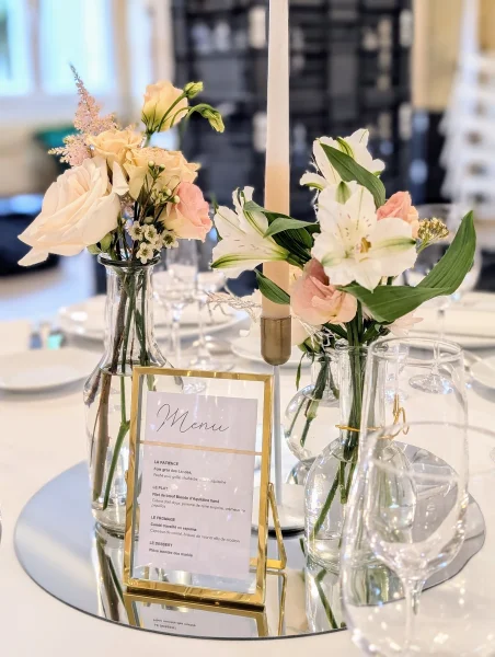 Centre de table de mariage classique avec roses, alstroemères, lisianthus dans des vases, bougie et menu sur miroir.