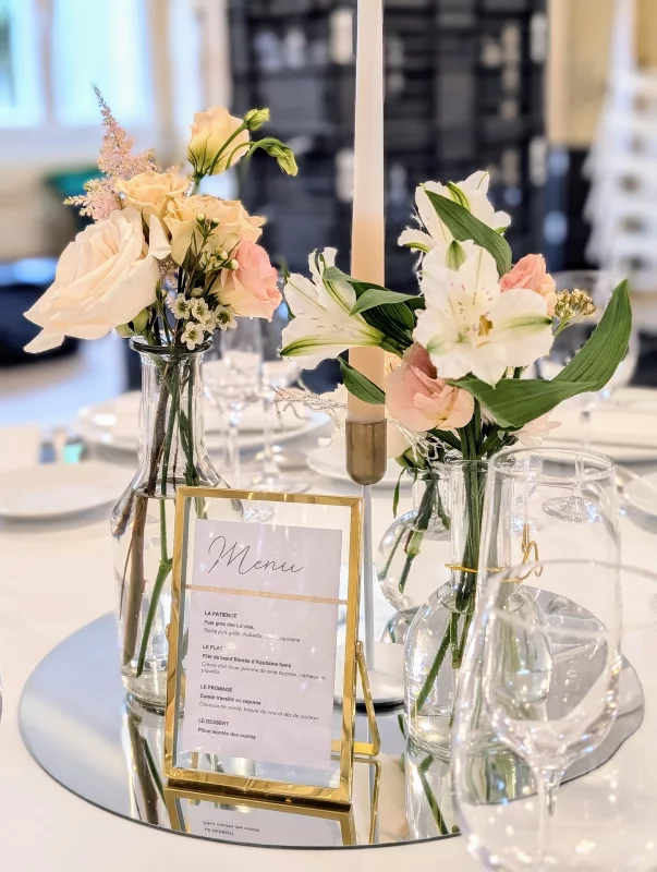 Centre de table de mariage classique avec roses, alstroemères, lisianthus dans des vases, bougie et menu sur miroir.