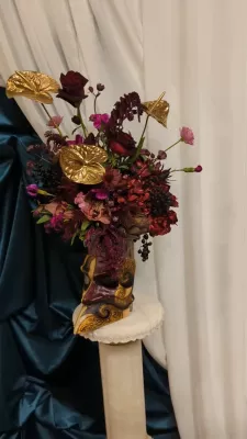Composition florale classique et élégante avec roses bordeaux, anthuriums dorés, et orchidées magenta dans un vase original.