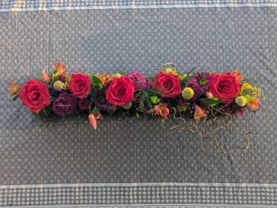 Centre de table allongé style bohème pour mariage, avec roses rouges, dahlias magenta et feuillage vert varié.