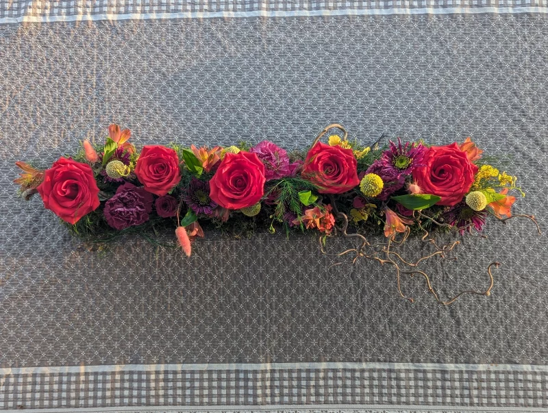 Elongated Bohemian-inspired table centrepiece for a wedding, featuring red roses, magenta dahlias, and varied green foliage.