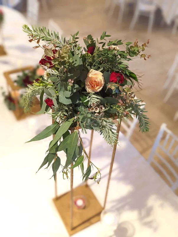 Romantic garden-style floral arrangement with peach and burgundy roses, eucalyptus and lush foliage on a tall wooden structure.