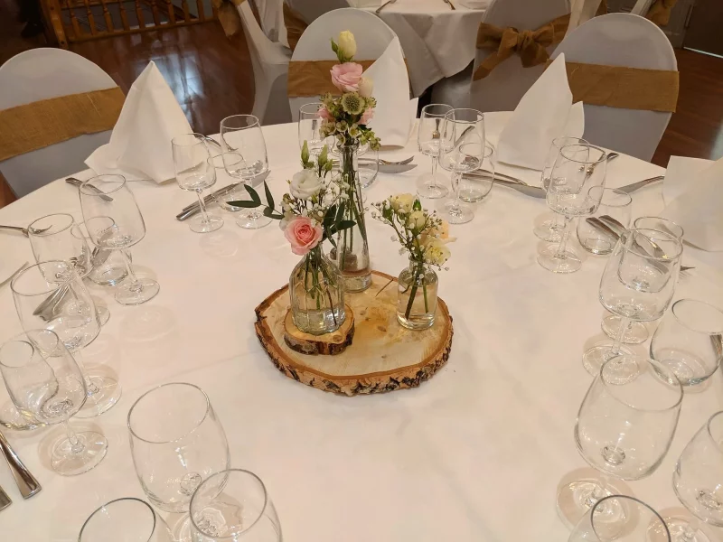 A rustic wedding table centrepiece featuring white and blush roses, eucalyptus, and a wooden log slice. A spring-inspired floral design.