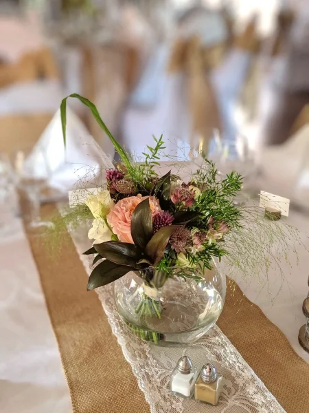 Centre de table champêtre avec roses pêche, feuillages variés et fleurs pourpres dans un vase boule en verre, mariage.