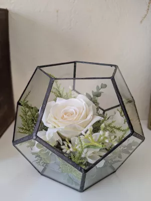 Composition florale de mariage moderne avec rose blanche, fougère et feuillage vert dans un terrarium géométrique en verre.