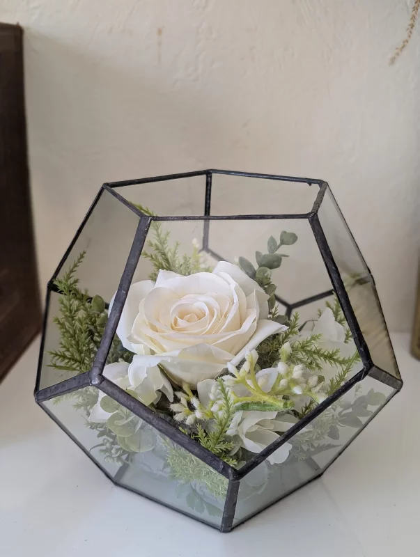 Composition florale de mariage moderne avec rose blanche, fougère et feuillage vert dans un terrarium géométrique en verre.