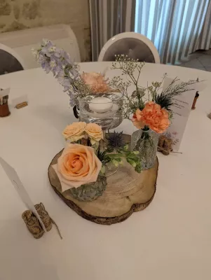 Centre de table champêtre avec rose pêche, œillet orange, delphinium bleu et gypsophile sur une tranche de bois.