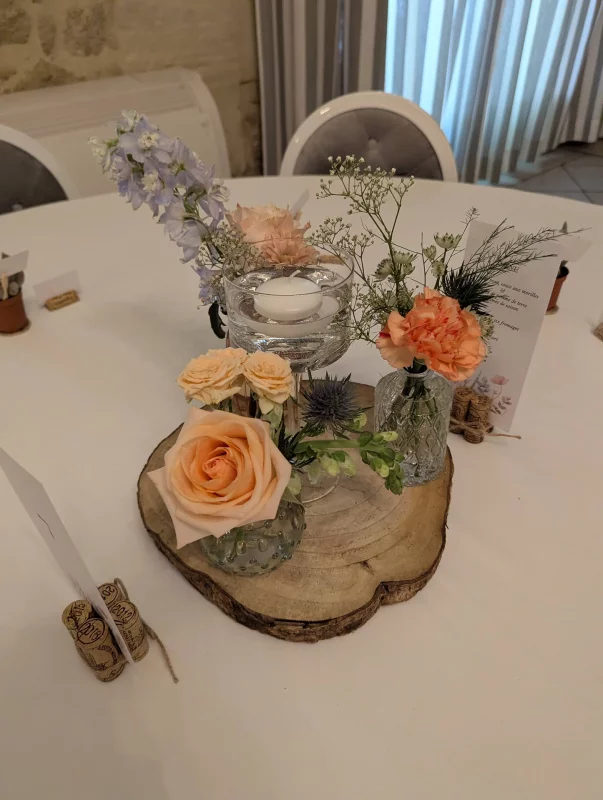 Centre de table champêtre avec rose pêche, œillet orange, delphinium bleu et gypsophile sur une tranche de bois.
