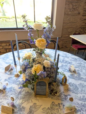 Centre de table classique pour mariage avec roses, hortensias bleus et delphiniums, sur nappe à motif Toile de Jouy.