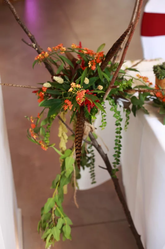 Composition scénographique bohème rustique avec branches, fleurs oranges et plumes.