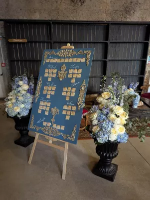 Grande composition florale classique sur colonne noire avec roses crème, hortensias bleus et delphiniums pour un mariage.